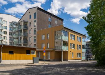 Puuterunkoinen ja vaaleasävyinen kerrostalokokonaisuus, jossa on lasitetut parvekkeet ja moderni julkisivu. Etualalla näkyy pihatie ja rakennuksen sisäänkäynti, taustalla sininen taivas ja pilviä.