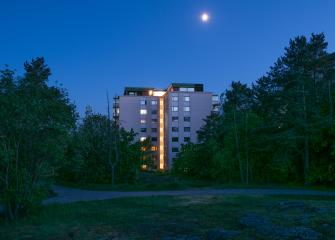Ilta- tai yöaikaan kuvattu näkymä metsäisen alueen halki kohti kerrostaloa, jonka keskiosa on valaistu. Taivaalla näkyy kirkas kuu ja rakennusta ympäröivät tummat puut.