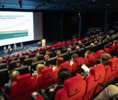 Kuva tapahtumapaikasta, jossa istuimia rivejä, etualalla iso näyttö ja lavalla neljä henkilöä istumassa paneelikeskustelijoina.