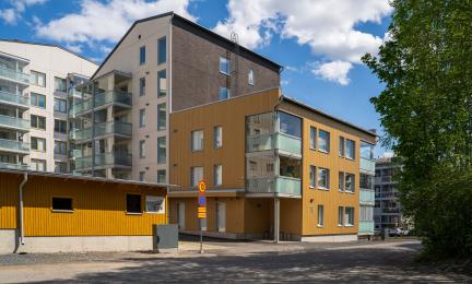 Puuterunkoinen ja vaaleasävyinen kerrostalokokonaisuus, jossa on lasitetut parvekkeet ja moderni julkisivu. Etualalla näkyy pihatie ja rakennuksen sisäänkäynti, taustalla sininen taivas ja pilviä.