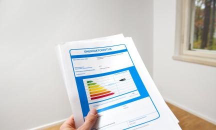 Kädessä pidetty energiatodistusasiakirja, jonka etusivulla näkyy värillinen energialuokituskaavio. Taustalla on vaalea, lähes tyhjä huone ja ikkuna oikealla.
