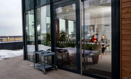 Lasiseinäinen huone, jossa kaksi iäkästä henkilöä istuu pöydän ääressä keskustelemassa; ulkopuolella näkyy terassi, lumisia kohtia ja kaupunkimaisema taustalla.