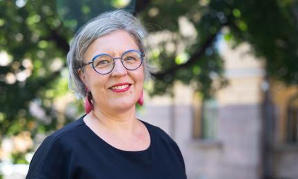 Sanna Hughes, Valtion tukeman asuntorakentamisen keskuksen johtaja, seisoo ulkona vihreiden puiden edessä ja katsoo kameraan hymyillen.