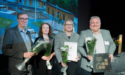 Neljä henkilöä seisoo lavalla palkintotilaisuudessa kukkakimppujen ja kunniakirjan kanssa. Taustalla on projisoitu kuva A‑Kruunun Suorannankatu 18 ‑asuntokohteesta.