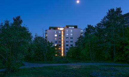 Ilta- tai yöaikaan kuvattu näkymä metsäisen alueen halki kohti kerrostaloa, jonka keskiosa on valaistu. Taivaalla näkyy kirkas kuu ja rakennusta ympäröivät tummat puut.
