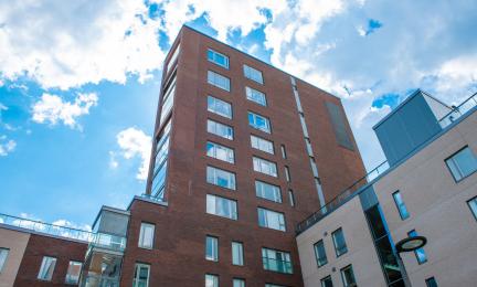 Punatiilinen kerrostalo kirkkaassa päivänvalossa, taustalla sininen taivas ja valkoisia pilviä. Rakennuksessa on useita ikkunoita ja sen vieressä näkyy osia muista moderneista rakennuksista.