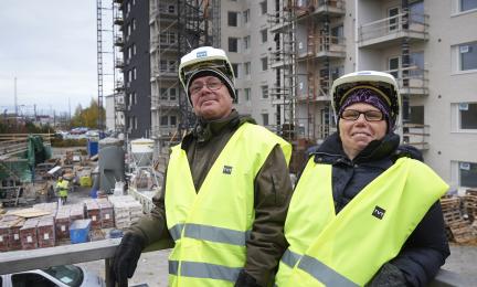 Kaksi henkilöä rakennustyömaalla, yllään keltaiset turvaliivit ja suojakypärät, taustalla kerrostalojen rakentaminen ja rakennusmateriaaleja.